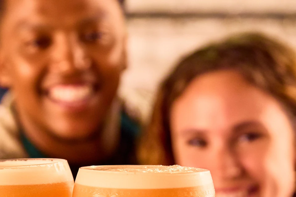 Two people smiling and holding up glasses filled with an orange beverage, with blurred background.