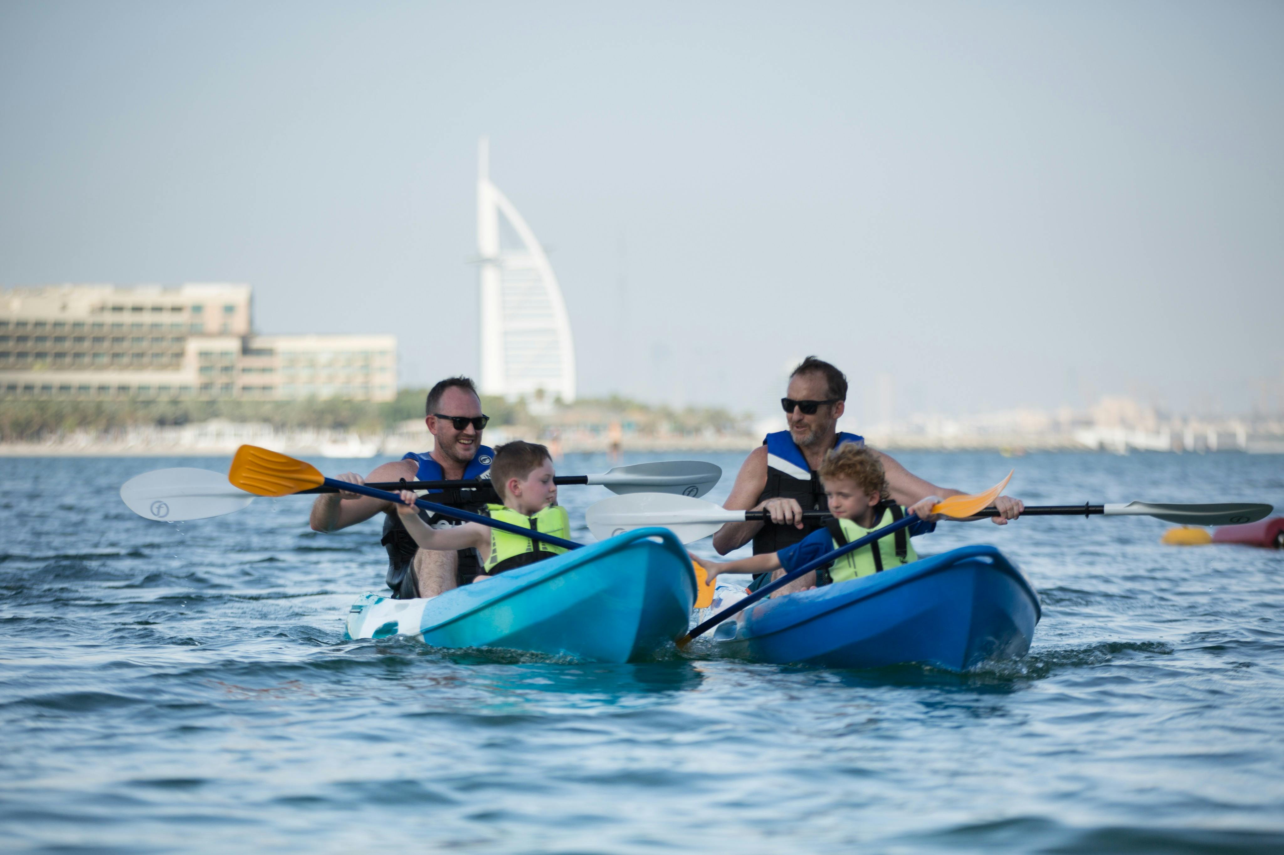 大人2人と子供2人が、大きな白い建物を背景に、穏やかな水の上で青いボートに乗ってカヤックをしています。