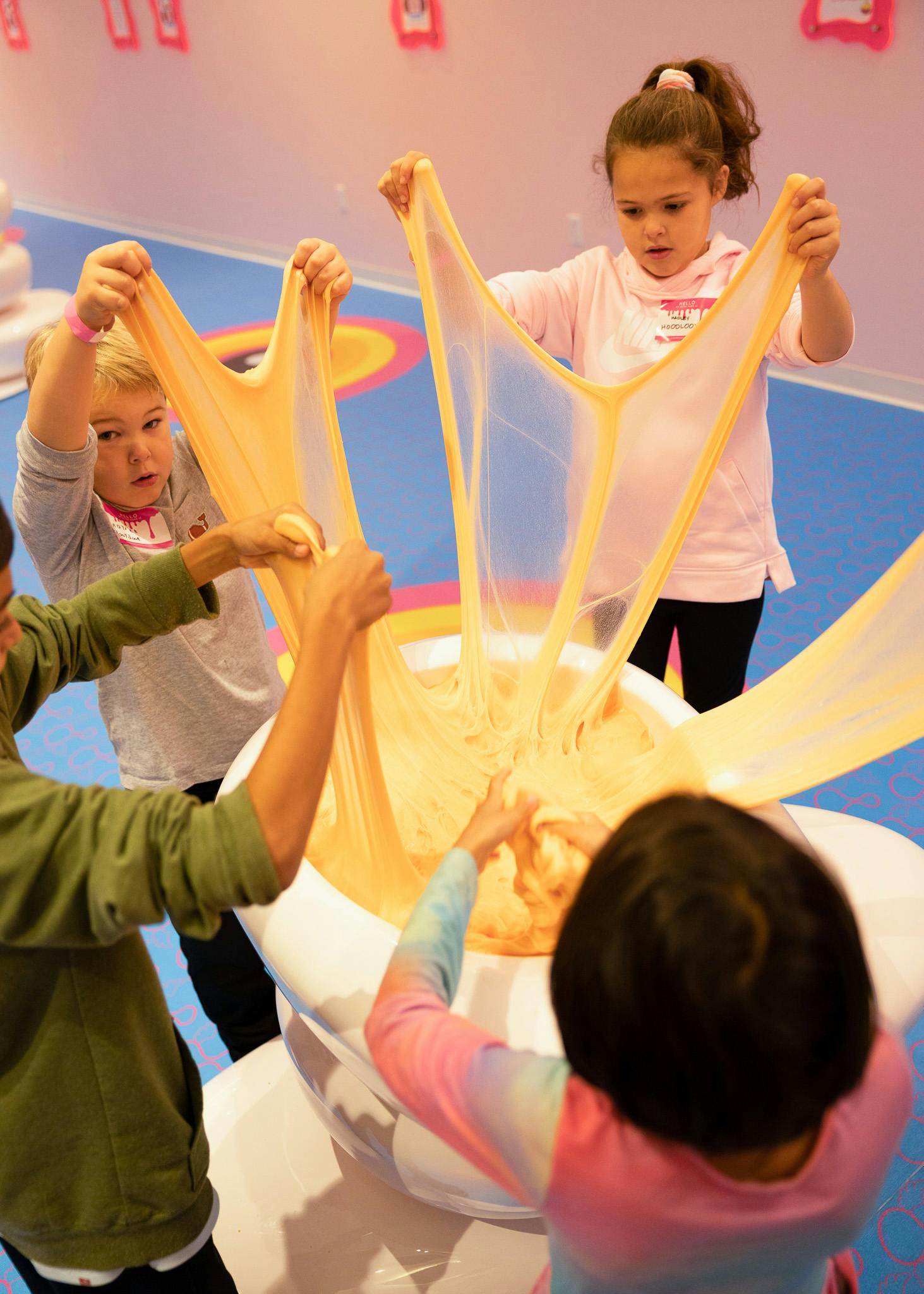 Fire børn strækker et stort parti slim på et legecenter med lyserød indretning og et blåt gulv.