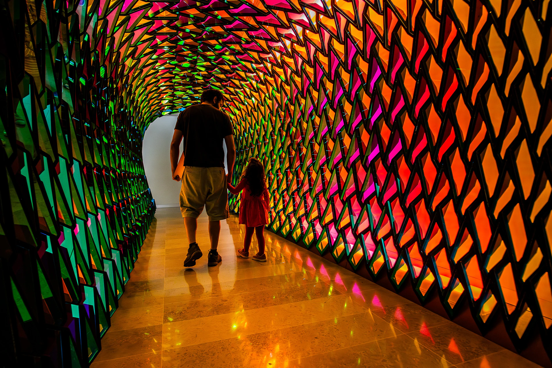 A man and a child walk through a tunnel with colorful, reflective geometric walls, illuminated by vibrant light patterns.