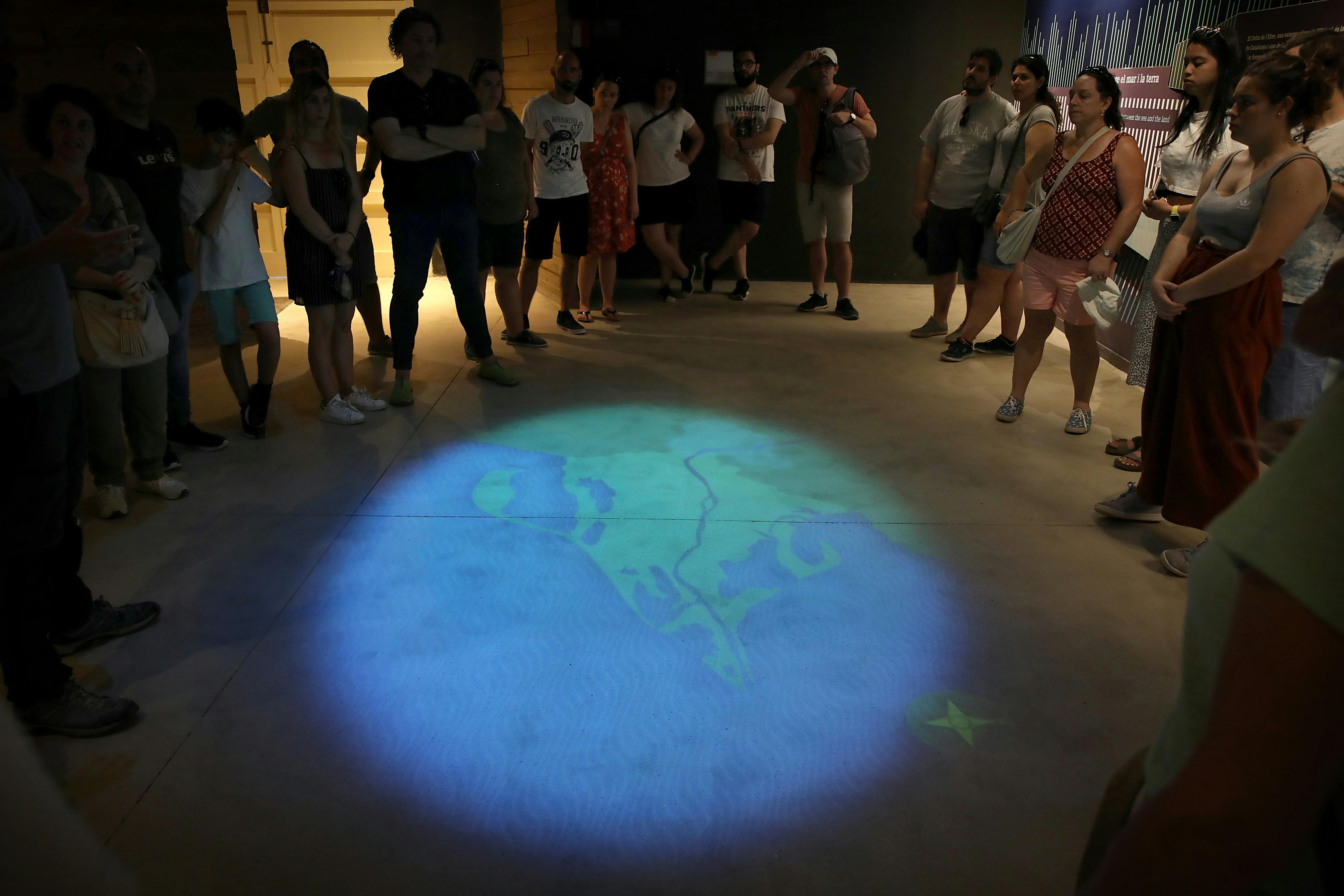 Μια ομάδα ανθρώπων στέκεται σε έναν κύκλο γύρω από έναν προβαλλόμενο χάρτη στο πάτωμα.