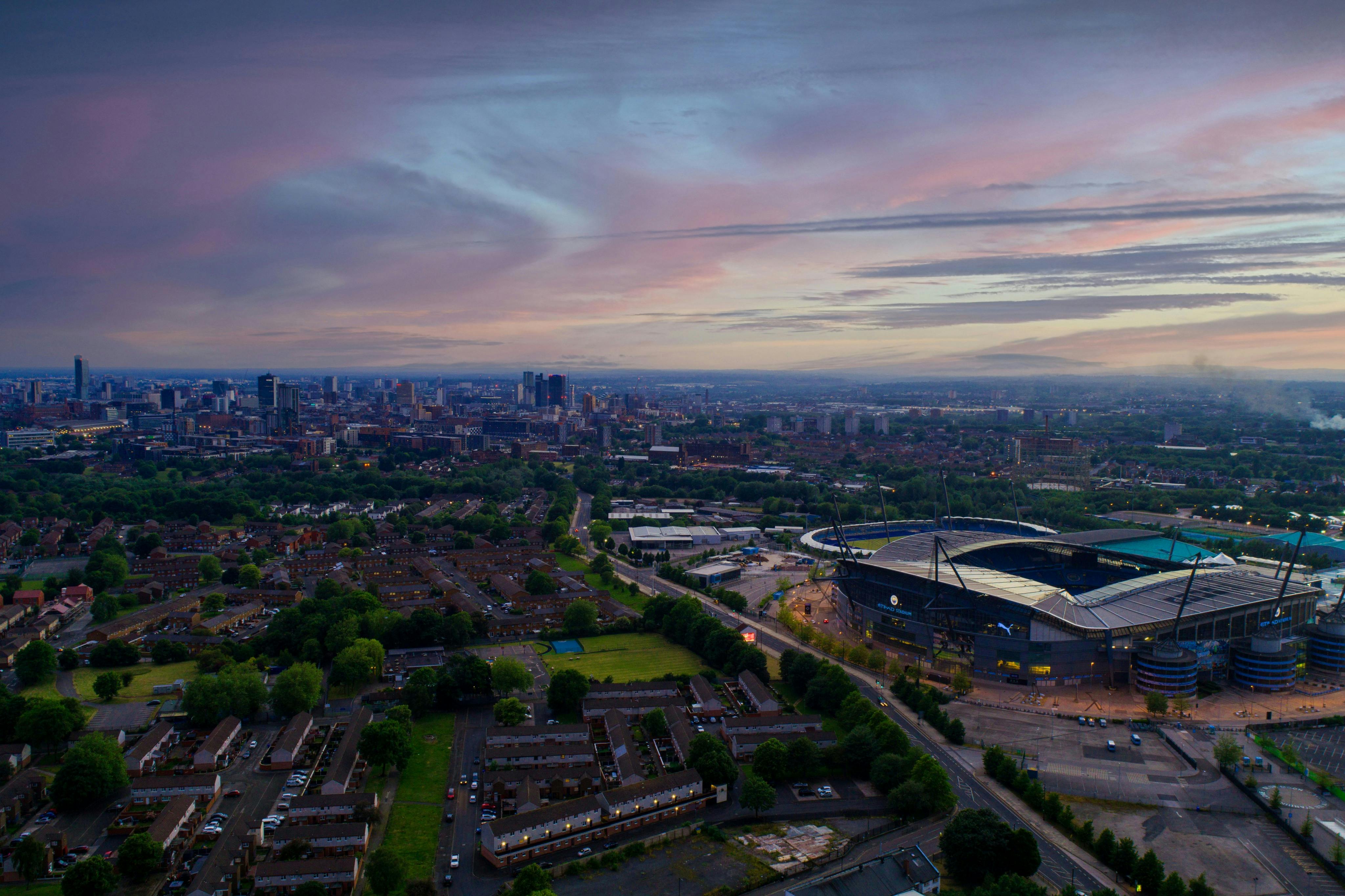 Vista aerea dello skyline di una città con un grande stadio, aree residenziali e spazi verdi sotto un cielo colorato e nuvoloso al tramonto.