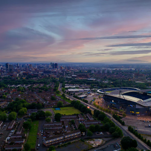 Flygfoto över en stadssilhuett med en stor stadion, bostadsområden och grönområden under en färgstark, molnig himmel i skymningen.