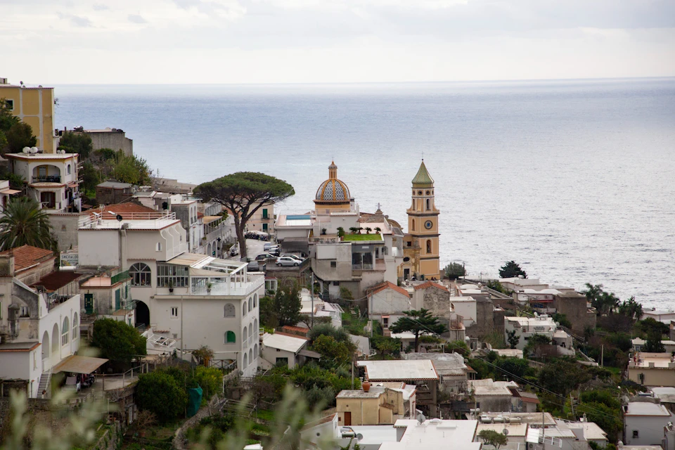 A coastal town with white buildings, a dome-shaped church, and a bell tower near the sea under a cloudy sky.