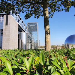 Modern byggnad med glas- och metallstruktur, stor tank, träd, frodig grönska och en reflekterande sfärisk struktur i bakgrunden.