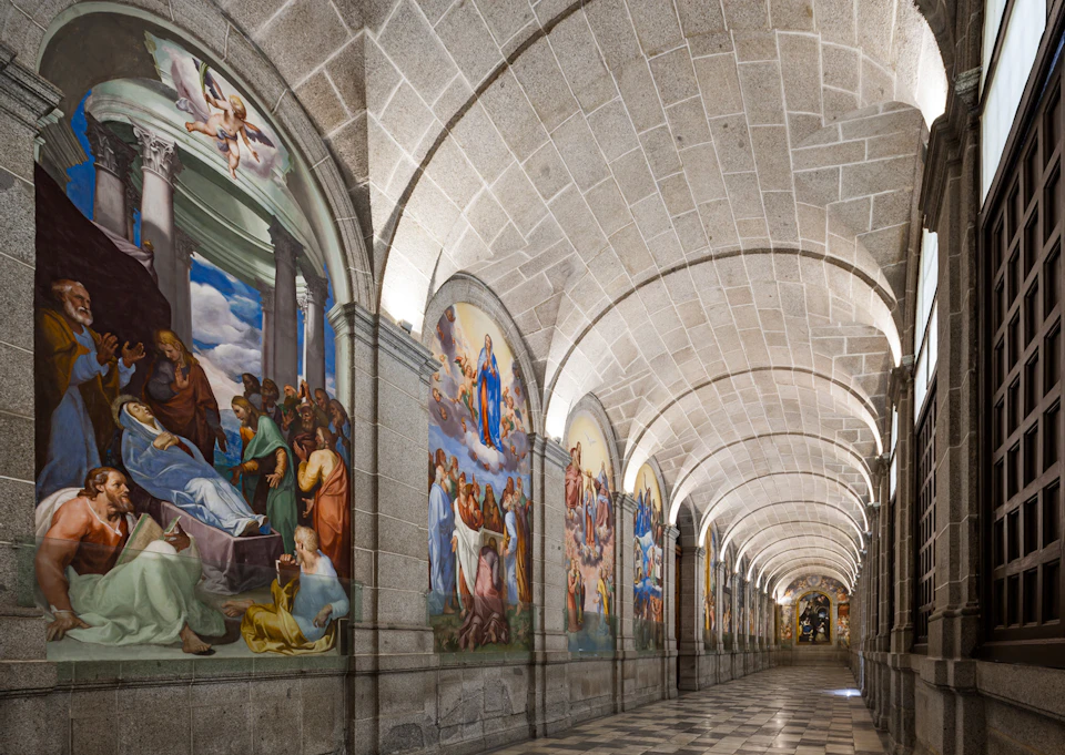 A long stone hallway with arched ceilings and large, colorful religious frescoes on the walls, depicting various biblical scenes.