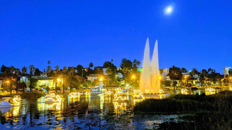 A brightly lit park at night with a large fountain, illuminated animal-shaped decorations on the water, and the moon in the sky.