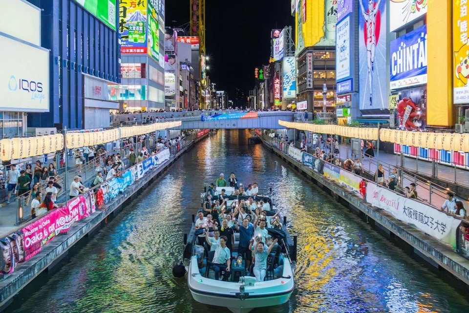 A boat with smiling passengers travels along a canal surrounded by bright neon signs and crowded sidewalks at night.