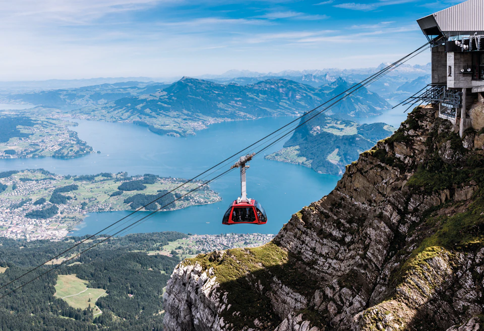 Red cable car traveling across a mountain landscape with a lake and distant hills under a blue sky.