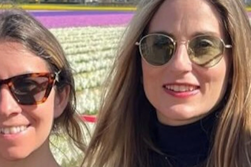 Three women wearing sunglasses stand in front of a field of colorful tulips under a partly cloudy sky.