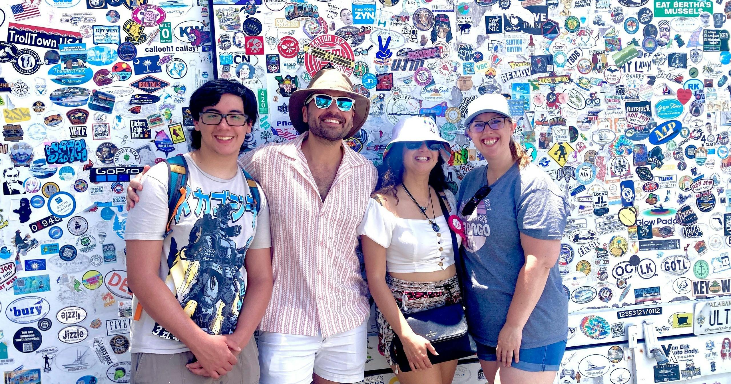 Trois personnes se tiennent devant un mur recouvert d'autocollants et sourient. L'une porte des lunettes, l'autre un chapeau et la troisième un chapeau blanc.