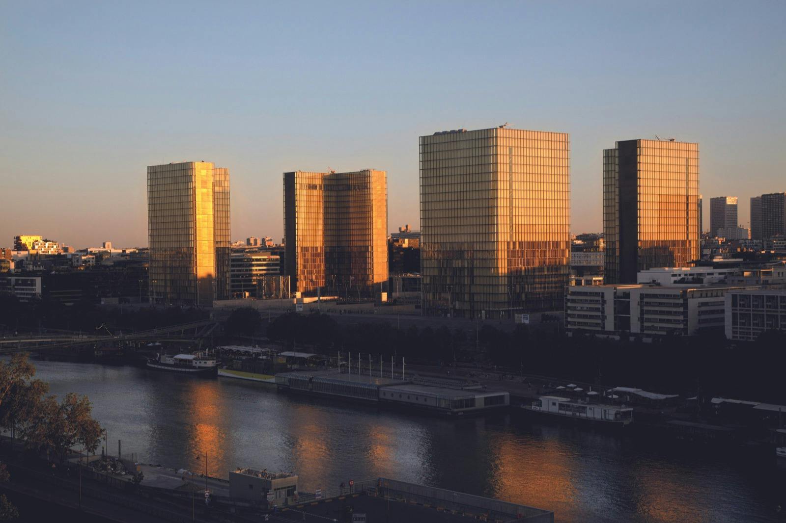 Fyra gyllene glastorn som reflekterar solljus nära en flod, med omgivande stadsbyggnader under en klar himmel.