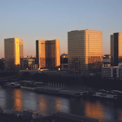 Fyra gyllene glastorn som reflekterar solljus nära en flod, med omgivande stadsbyggnader under en klar himmel.