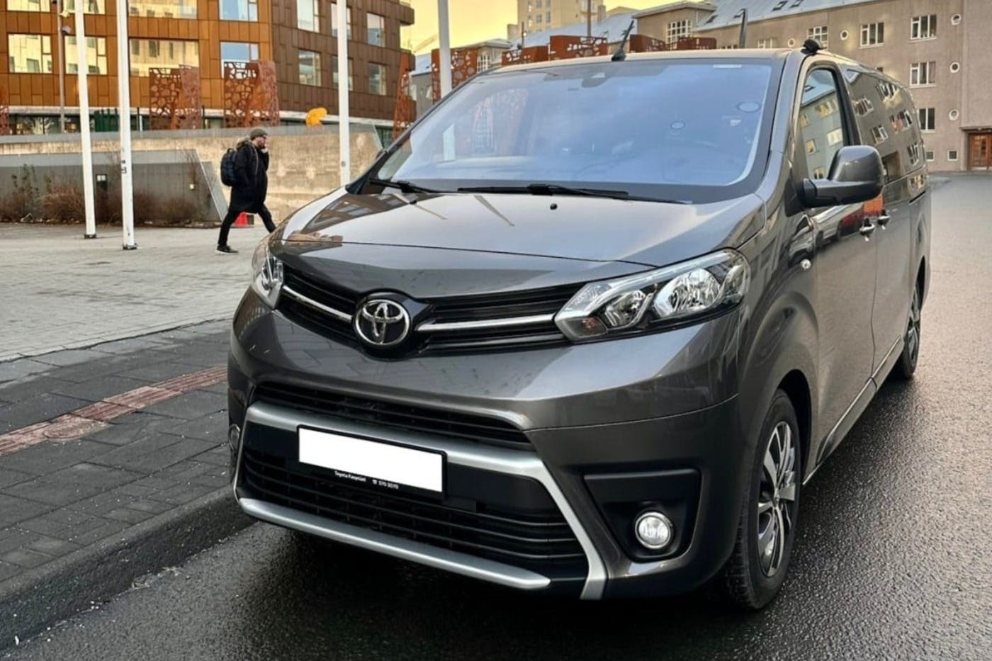 A gray Toyota minivan parked on a city street with buildings and a pedestrian in the background.