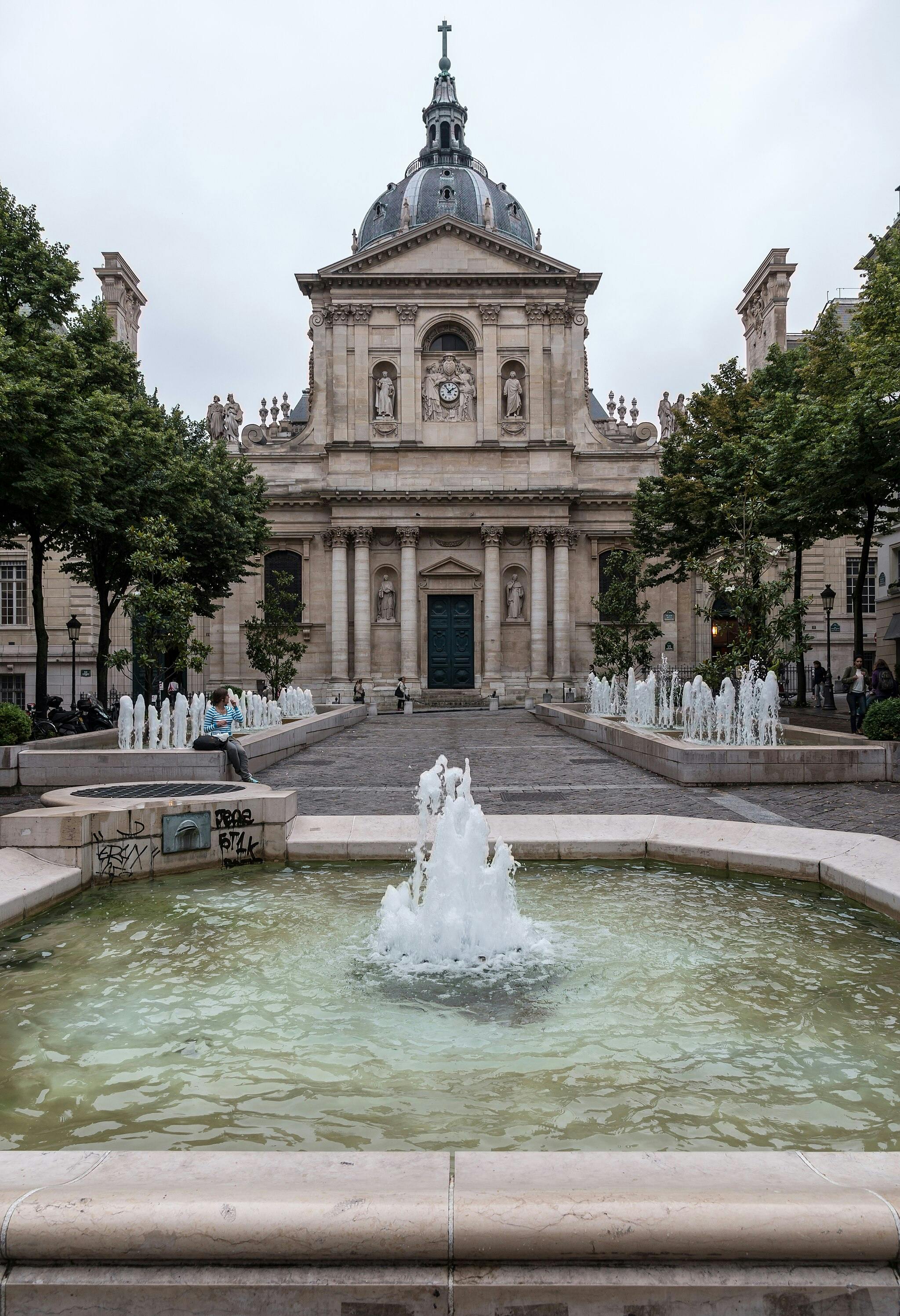 A stone building with statues and a clock, surrounded by trees and fountains, viewed across a pond with a central water spout.