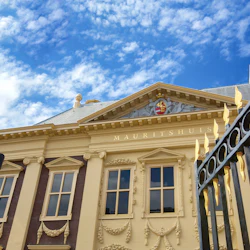 Elegant byggnadsfasad märkt "MAURITSHUIS" med utsmyckade fönster och en dekorativ grind toppad med gulddetaljer under en blå himmel.