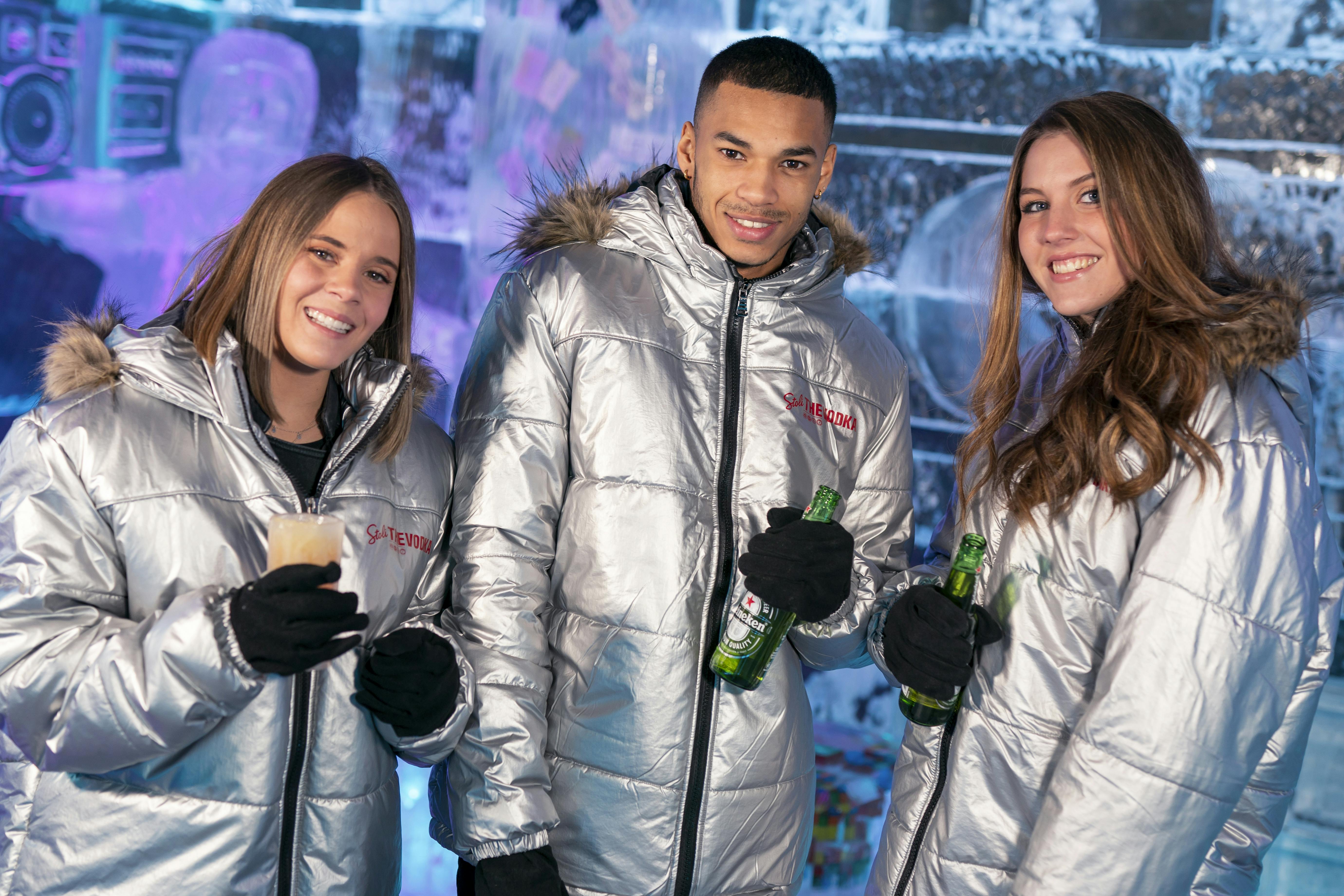 三个穿着银色夹克的人紧紧地站在一起，拿着饮料，微笑着，冰雪覆盖的背景。