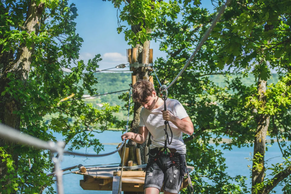 A person wearing a harness navigates a rope course in a lush, leafy tree canopy with a lake and hills in the background.