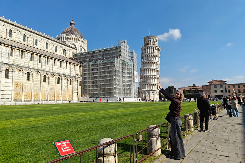 A person poses near the Leaning Tower of Pisa on a sunny day, with the tower and adjacent buildings in view.