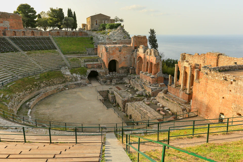 Ancient stone amphitheater with partial ruins, green seating areas, and a distant view of the sea. Trees and a small building in the background.