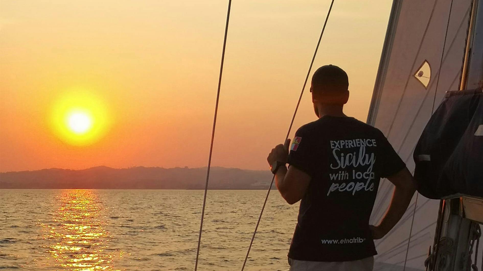Osoba v košili „Zažij Sicílii s místními lidmi“ stojící na lodi při západu slunce, držící provaz, s vodou a horami v pozadí.