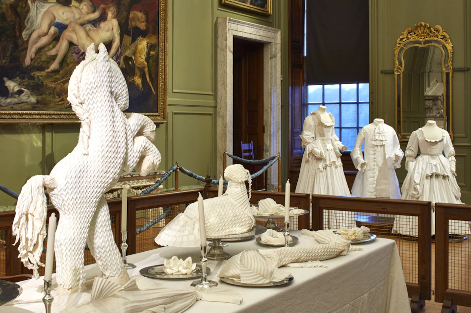 An ornate room with a table set with intricately folded white napkins and gowns displayed on mannequins in the background.