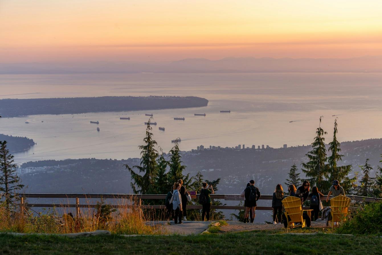 People enjoying a scenic view of a bay, ships, and a city skyline at sunset, with trees and chairs in the foreground.
