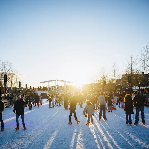 Tre barn skridskor; en skjuter en annan i en stol, under strängljus, med en stadsbakgrund och åkare i bakgrunden.