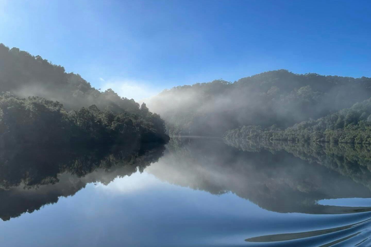 A serene lake surrounded by forested hills with mist and a clear blue sky reflected in the calm water.