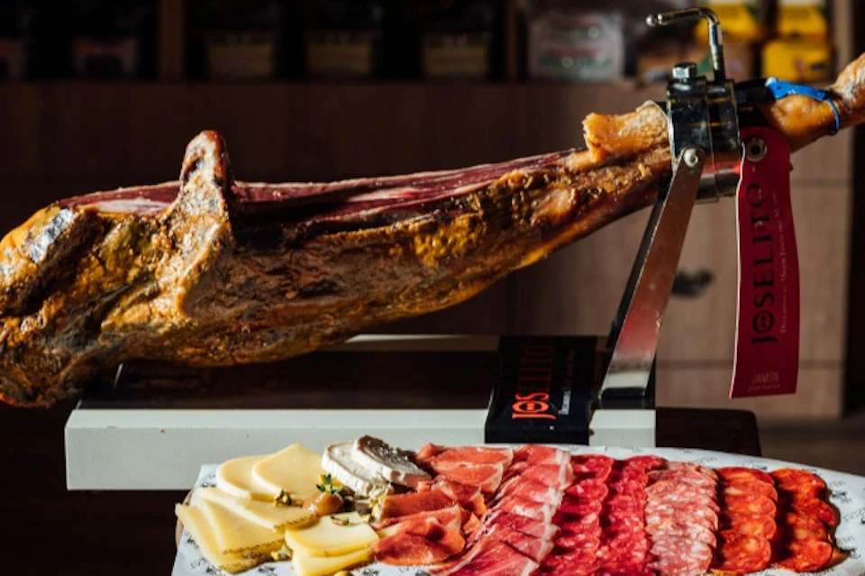 A cured ham leg on a metal stand with an assortment of sliced cheeses and meats displayed in the foreground.