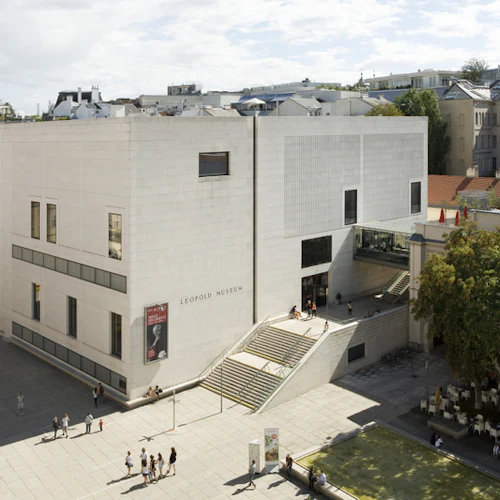 Stadsmiljö med en modern rektangulär byggnad märkt "Leopold Museum", promenader, träd och uteserveringar.