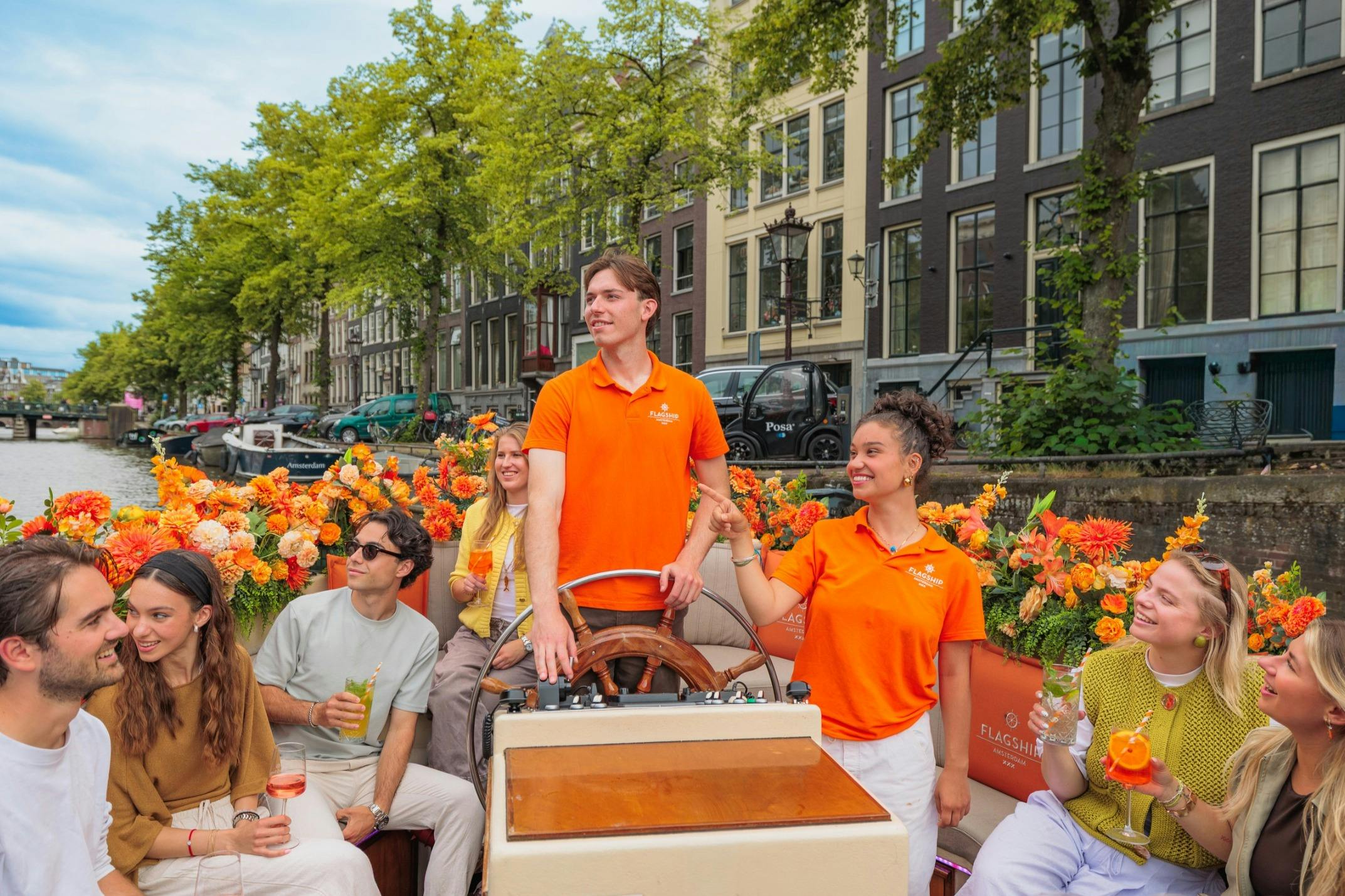 A group of people in a boat on a canal surrounded by flowers. Two individuals in orange shirts are steering the boat.