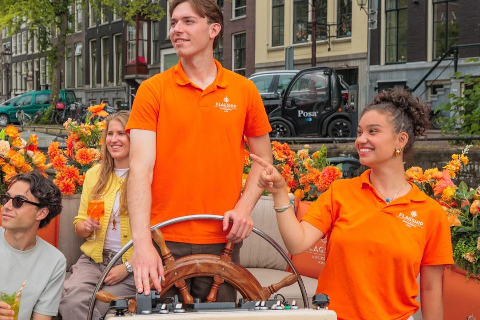 A group of people in a boat on a canal surrounded by flowers. Two individuals in orange shirts are steering the boat.
