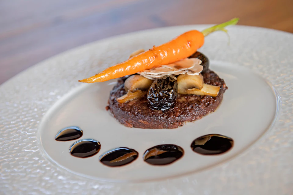 A plated gourmet dish features a roasted carrot on top of a patty with mushrooms, garnished with sliced almonds and balsamic dots.