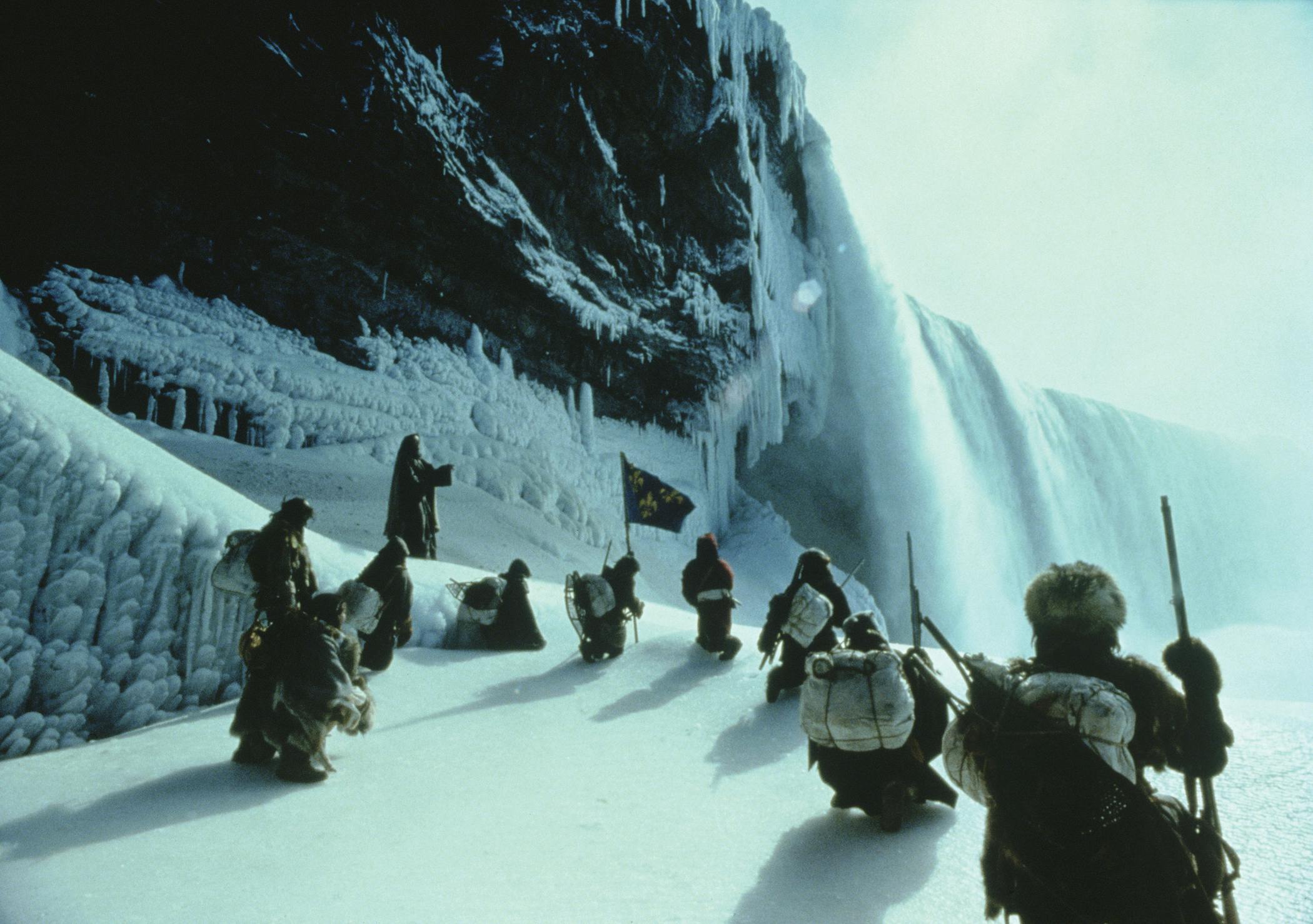 겨울 옷을 입은 사람들은 폭포가 있는 크고 수직인 얼음층 근처의 얼음 지형에 무릎을 꿇고 서 있습니다. 한 사람이 깃발을 들고 있습니다.