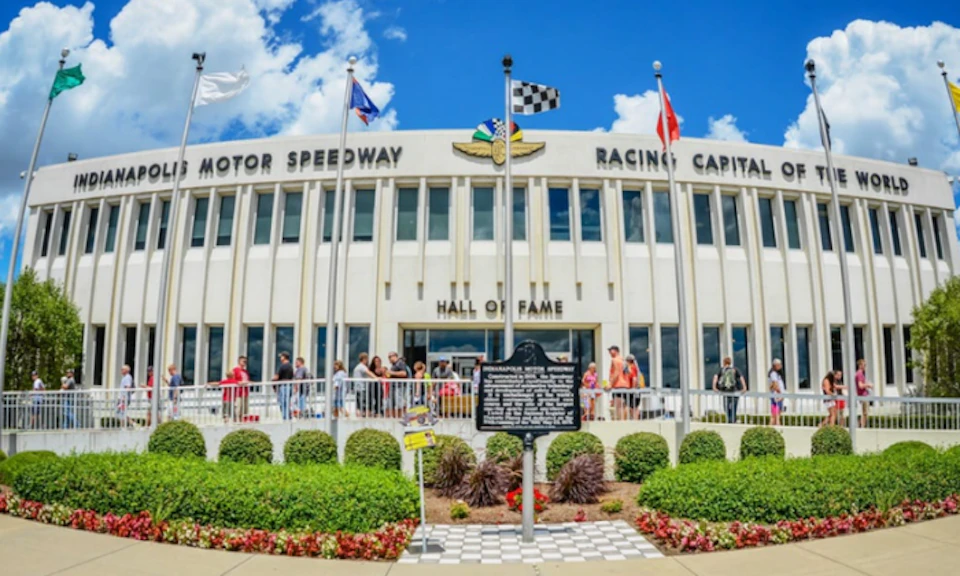 People stand outside a building labeled "Hall of Fame" at the Motor Speedway Racing Capital. Three flags fly above the entrance.