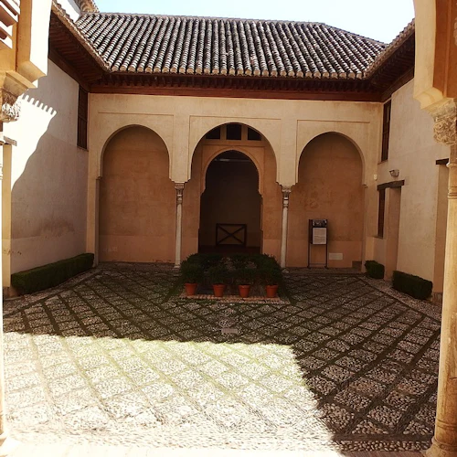 En solbelyst innergård med tegeltak, välvda dörröppningar, stenmosaikgolv och krukväxter i mitten.