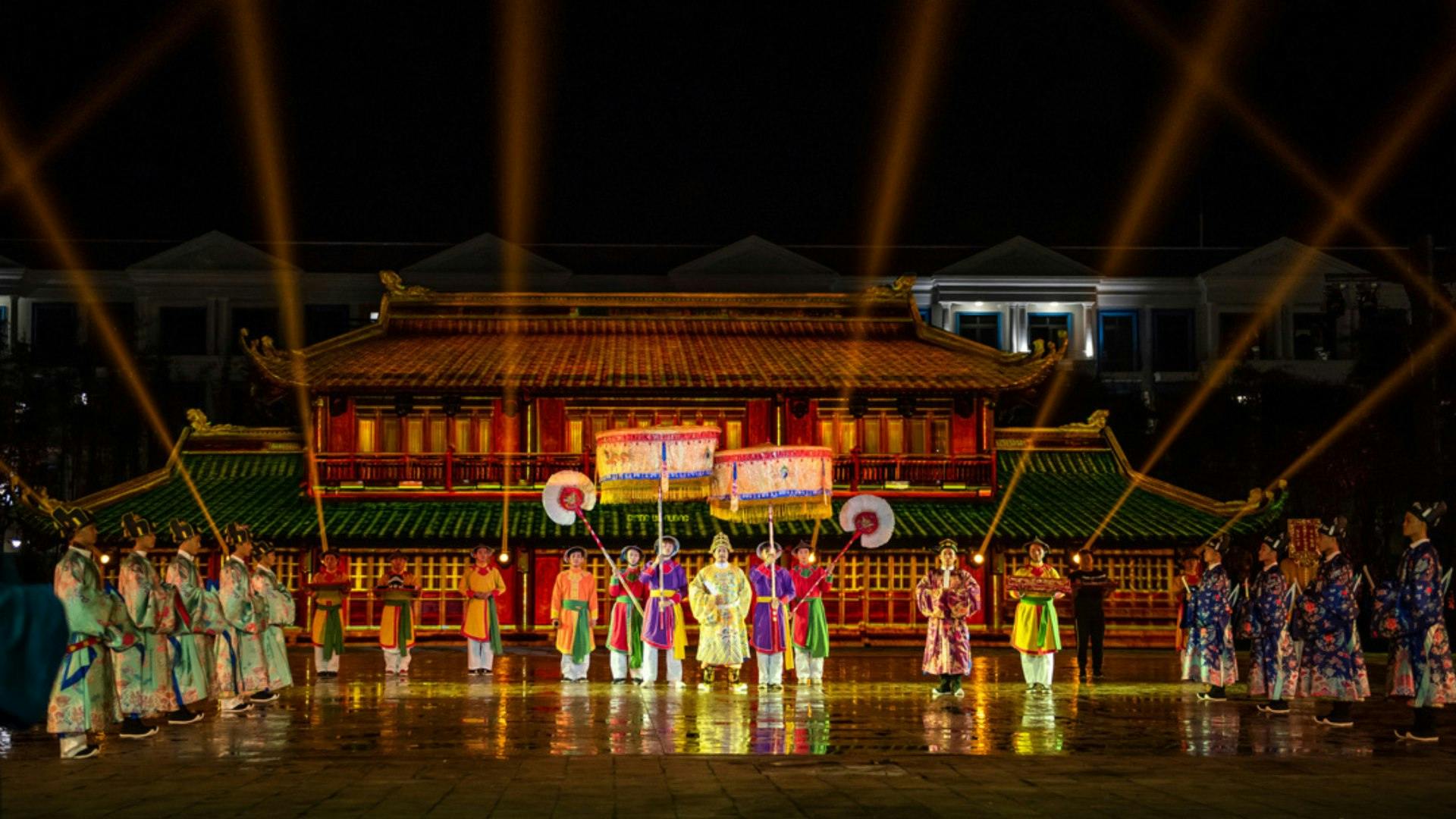 Performers in traditional attire with large fans and banners in front of a lit-up traditional building at night.