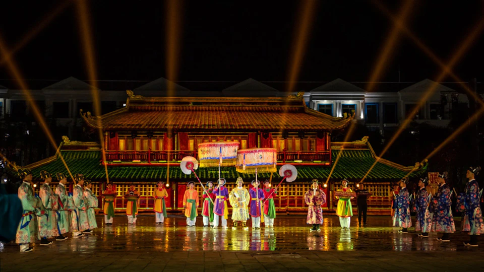 Performers in traditional attire with large fans and banners in front of a lit-up traditional building at night.