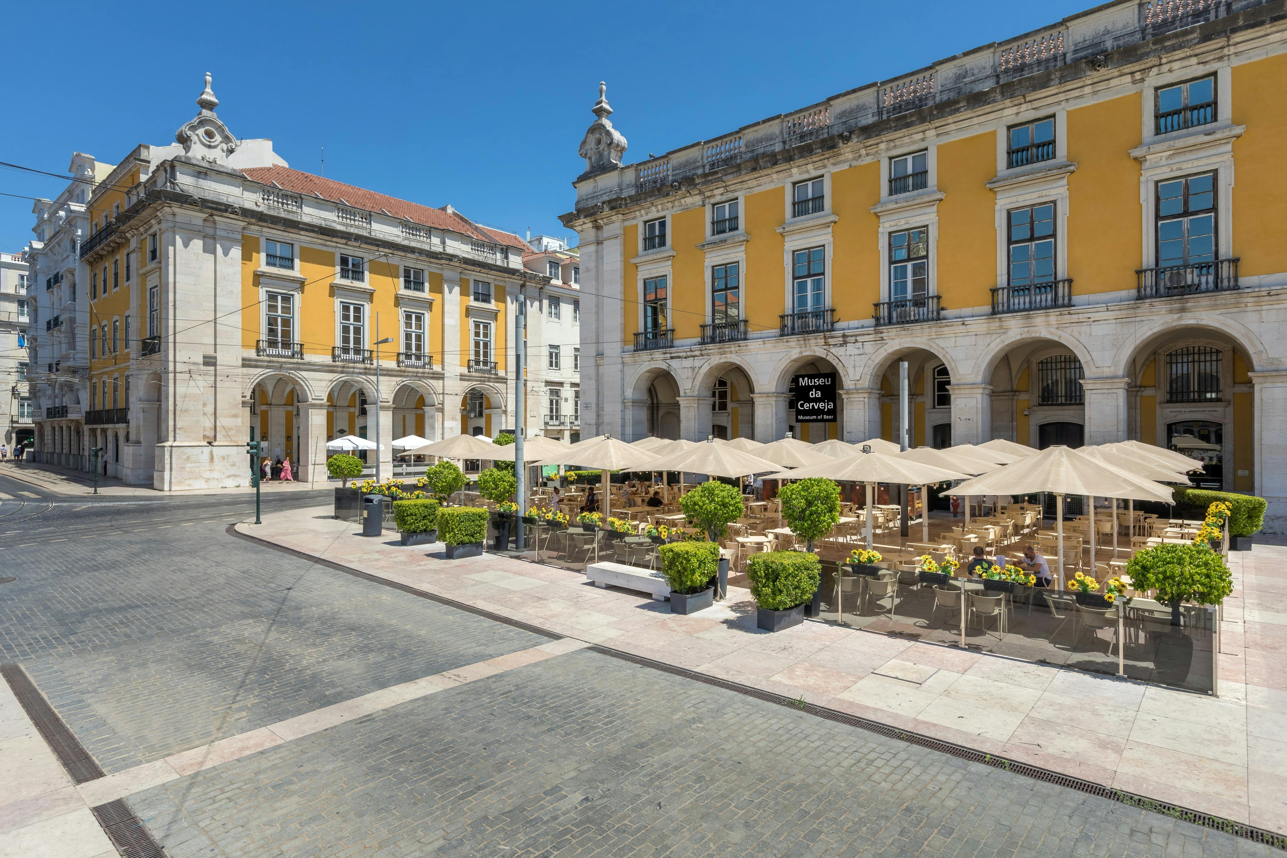 Ett torg med uteservering, gula byggnader och Museu da Cerveja-skylten under en klarblå himmel.