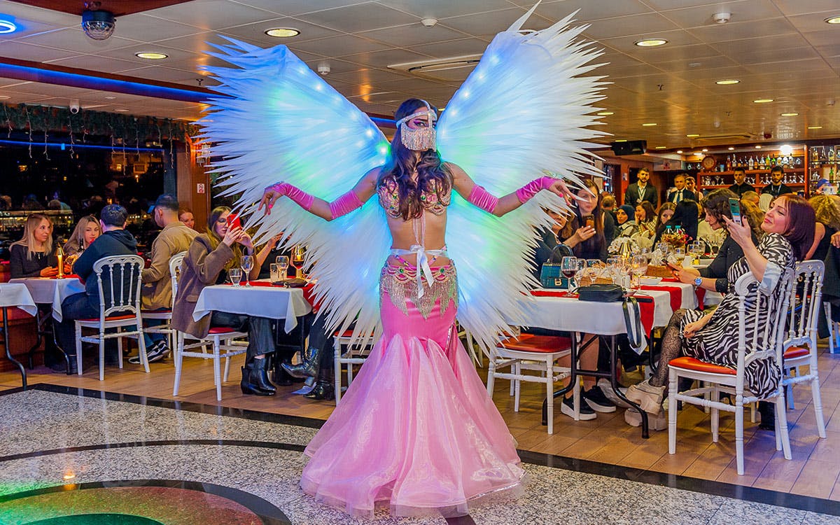 A performer in a pink and white costume with large illuminated wings dances in a restaurant while guests dine and watch.