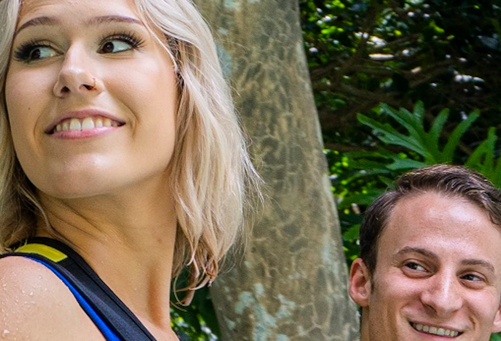 Two people in wetsuits stand in shallow water, surrounded by lush tropical plants and trees.