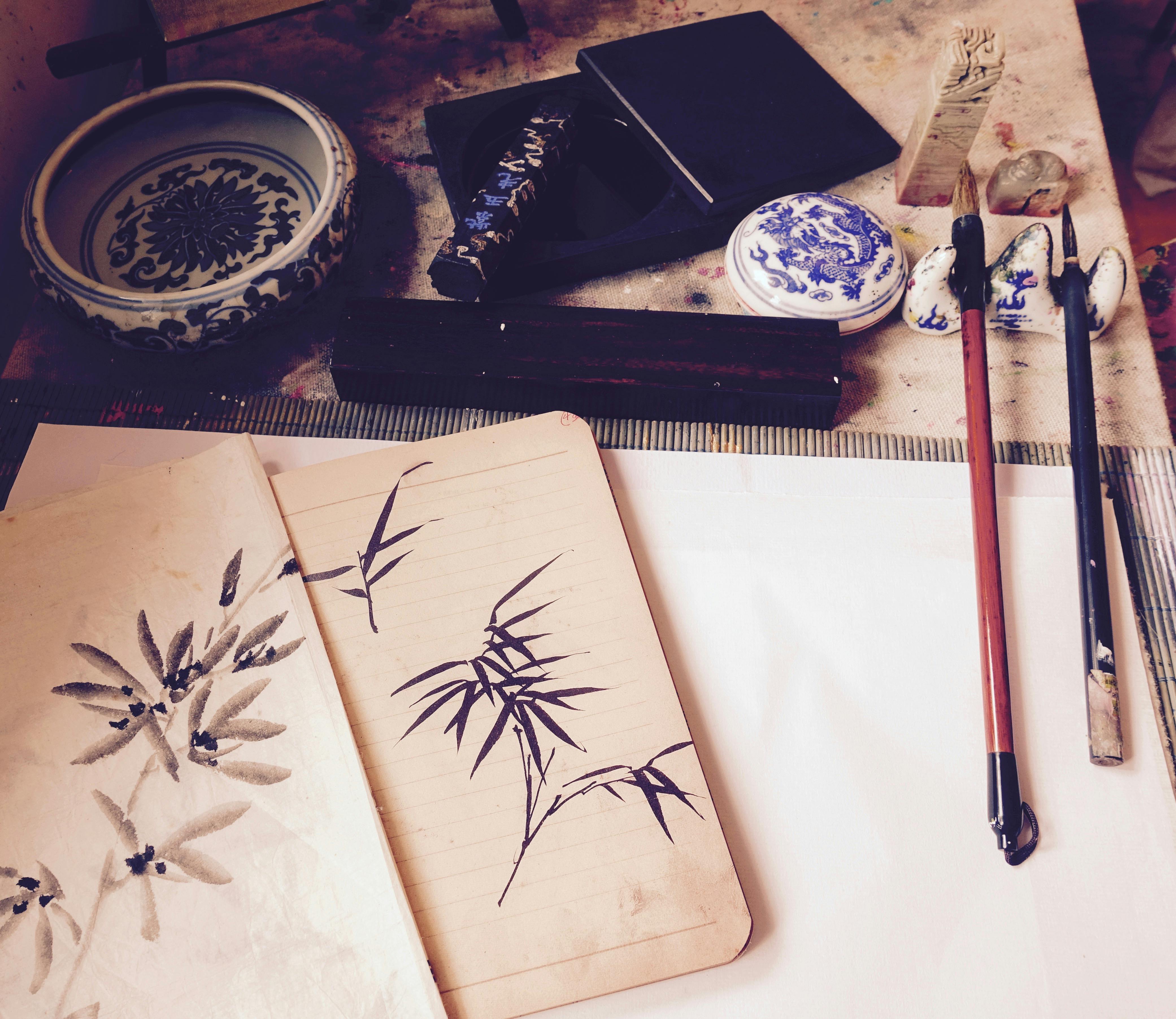 Traditional Chinese painting setup with brushes, ink stones, ceramics, and a notebook displaying bamboo and leaf sketches.