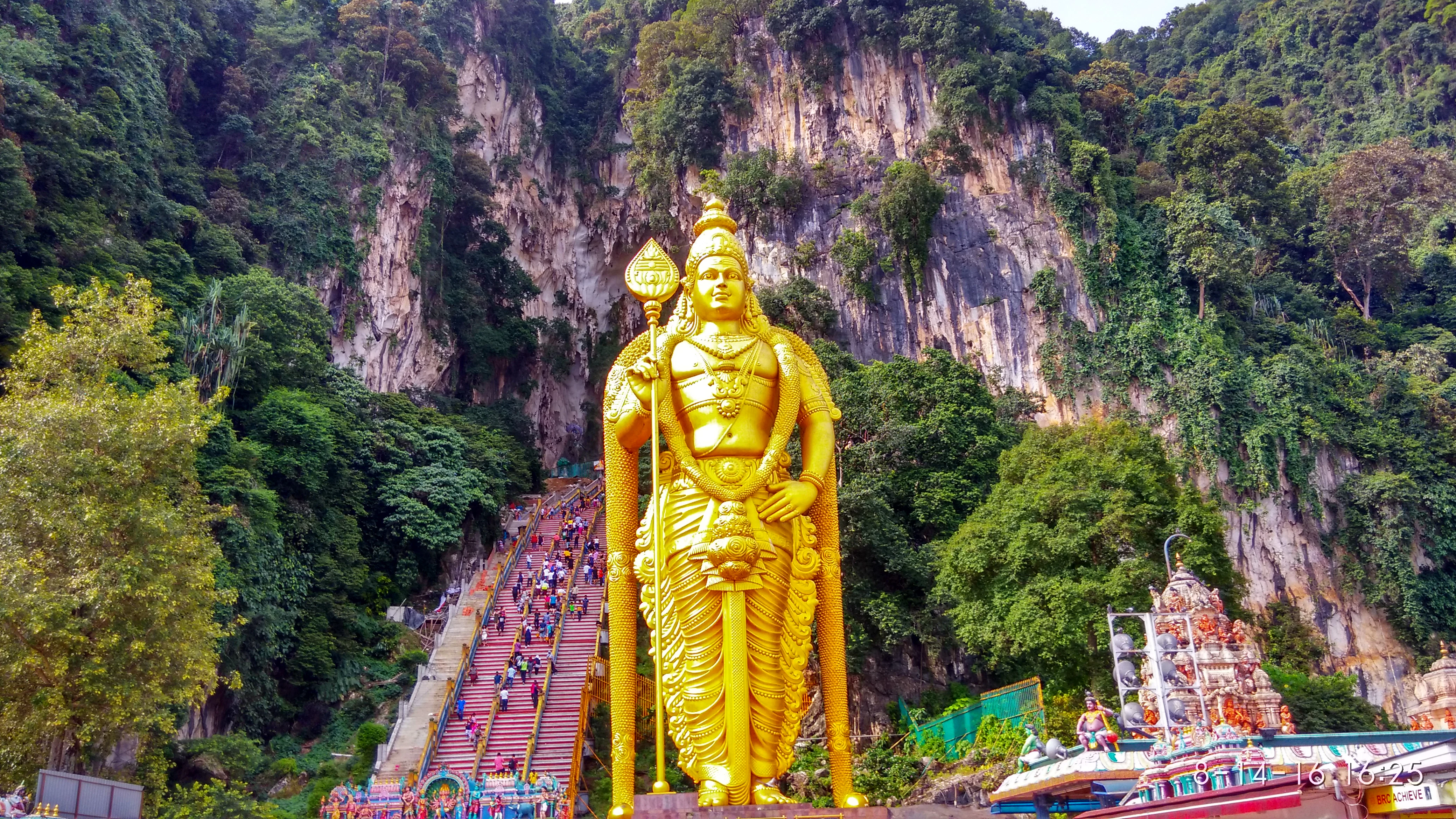 Golden statue of a deity stands at the base of steep, colorful stairs with people ascending, surrounded by lush greenery and rocky cliffs.