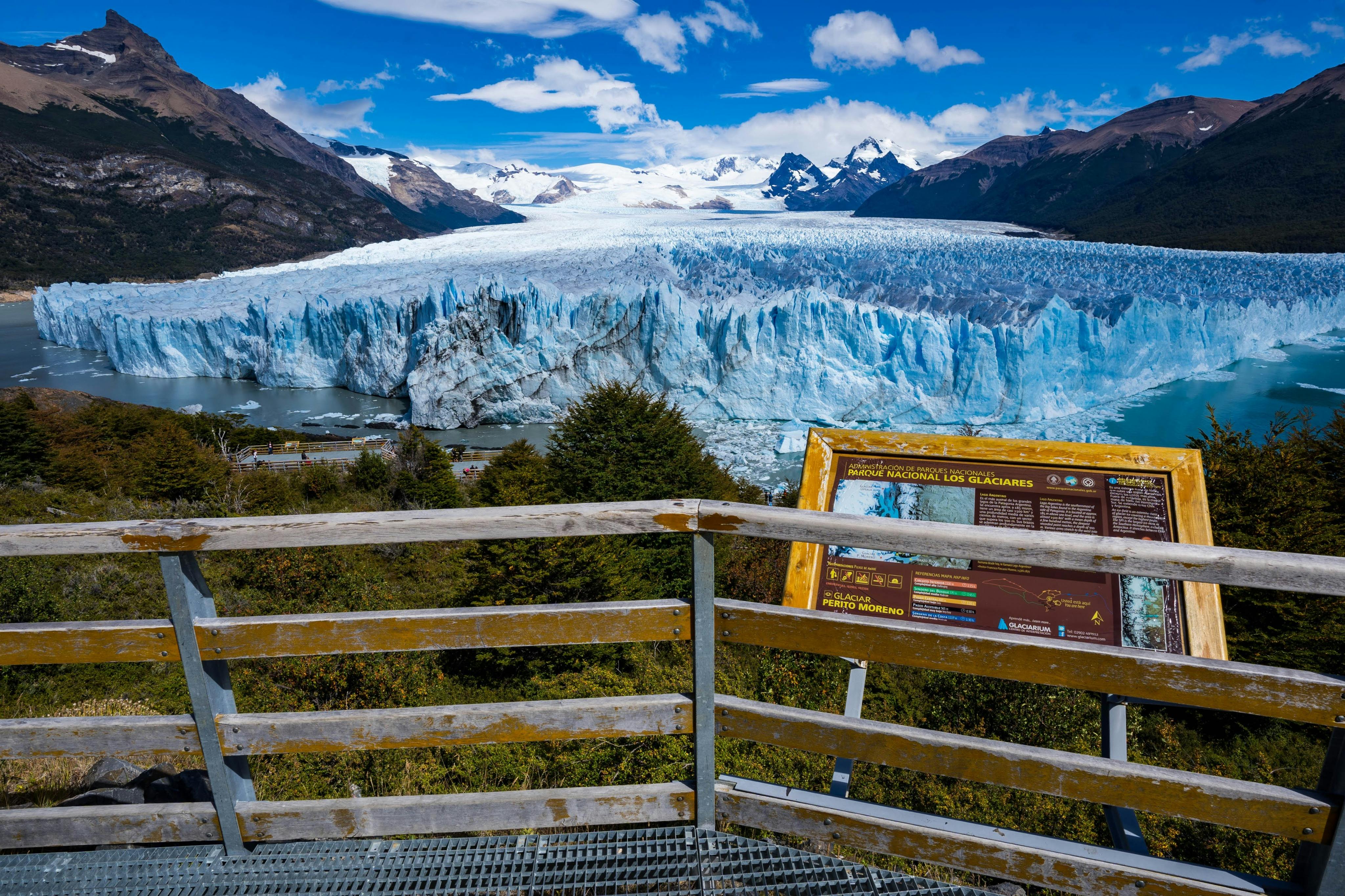 Wooden railing overlooking a glacier under a blue sky, with a signboard displaying information in the foreground.