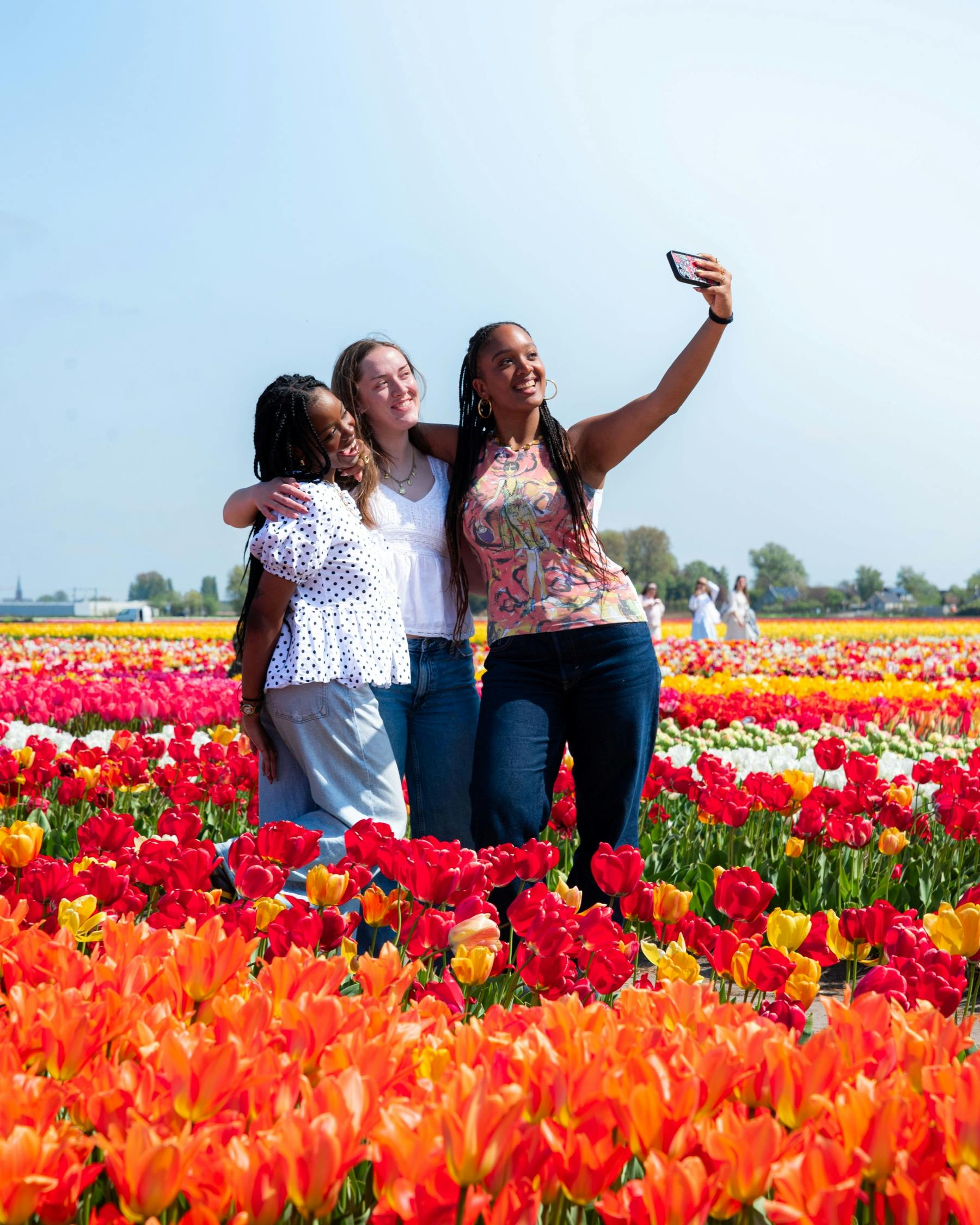 Three people standing in a field of colorful tulips, smiling and taking a selfie under a clear blue sky.