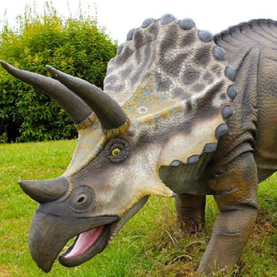 Model of a triceratops with detailed texture, showing its three horns and frill, positioned on grass with shrubbery in the background.