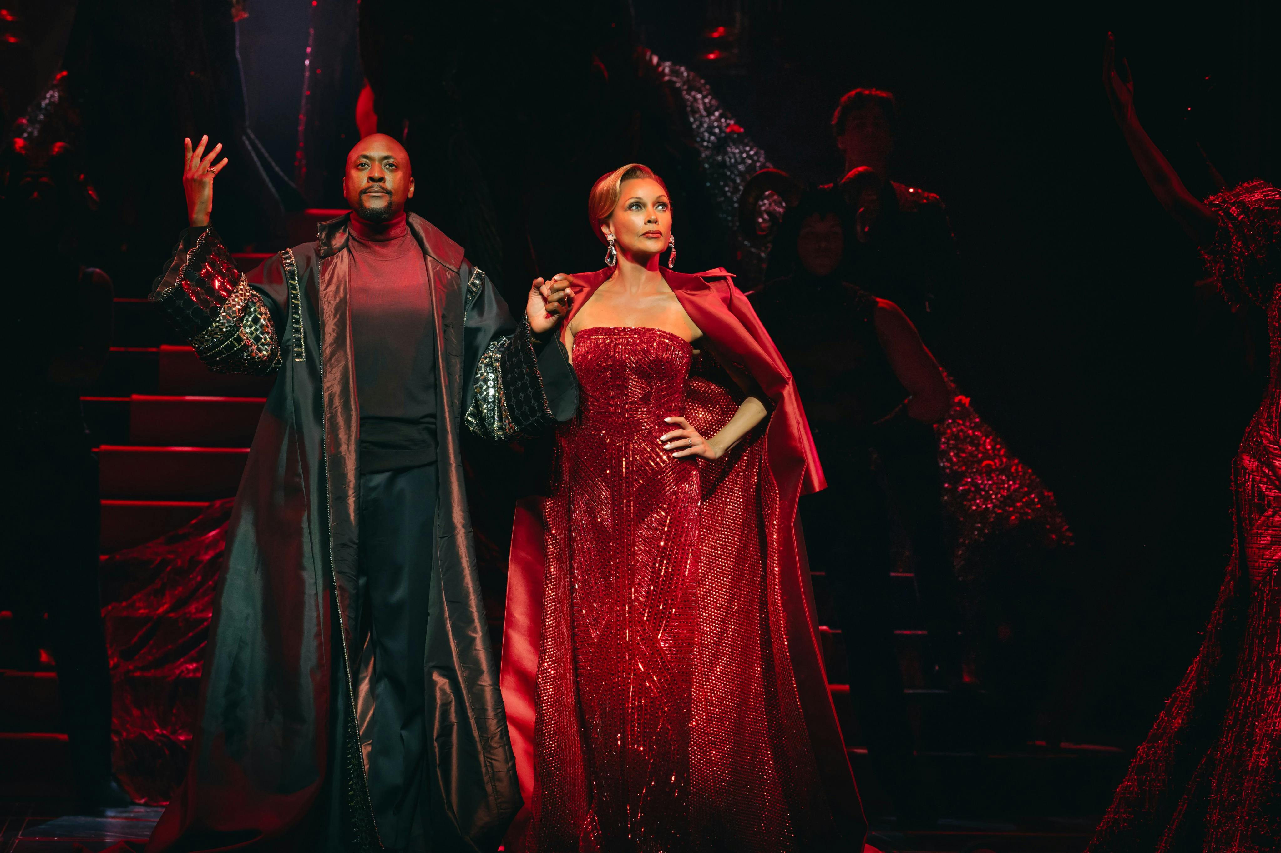 Two individuals stand in dramatic, elegant attire under red lighting. One wears a black coat; the other, a sparkling red gown.