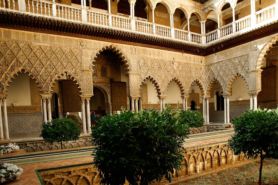 A historic courtyard with ornate arches, detailed carvings, greenery, and a water feature, surrounded by a two-story arcade.