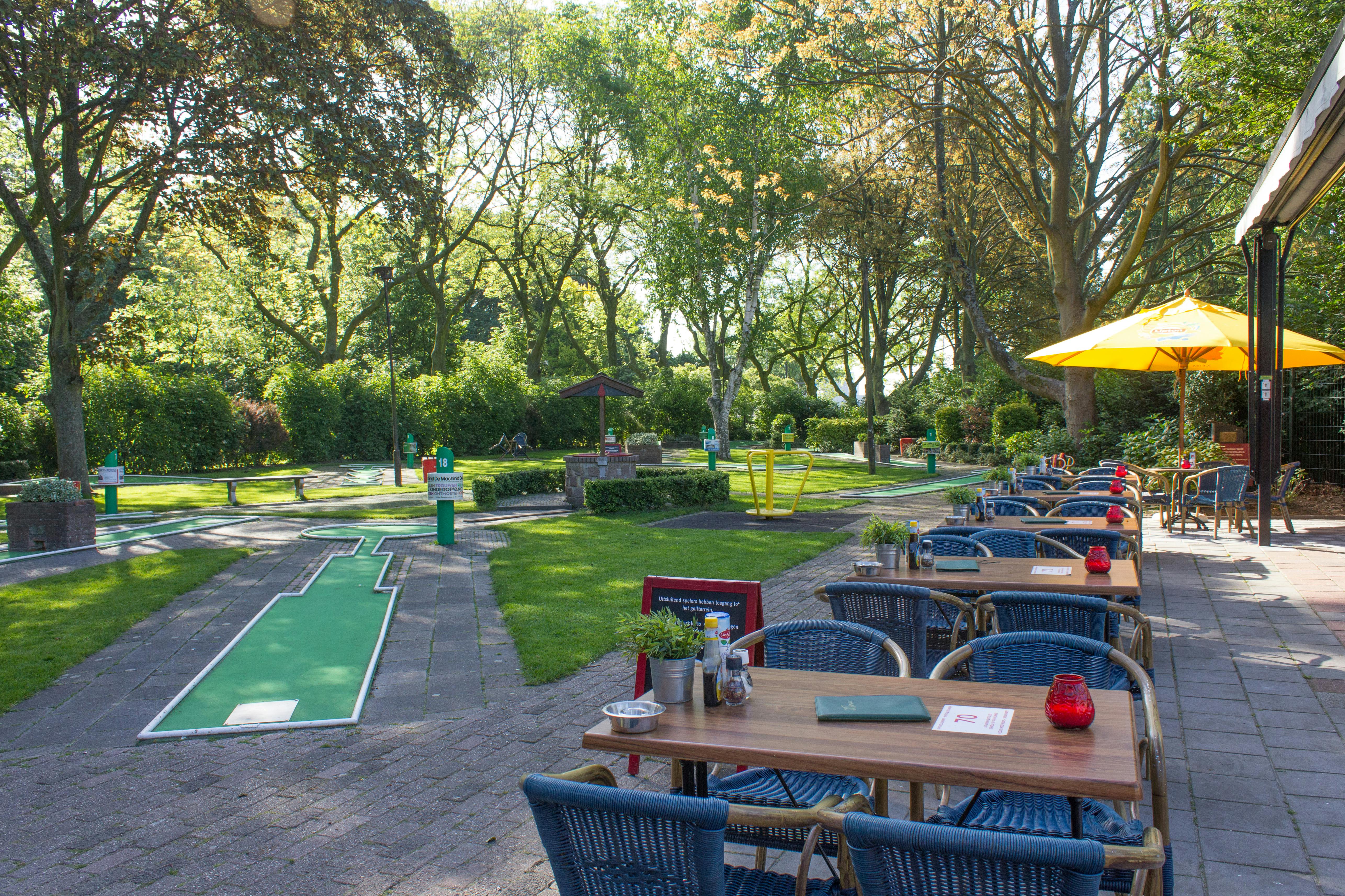 户外咖啡馆，迷你高尔夫球场旁边有蓝色柳条椅和木桌，四周环绕着郁郁葱葱的绿树和公园风光。